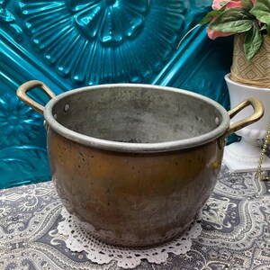 May include: A vintage copper pot with brass handles. The pot has a rounded body and a silver rim. The exterior has a warm, aged patina. The pot is sitting on a white lace doily.