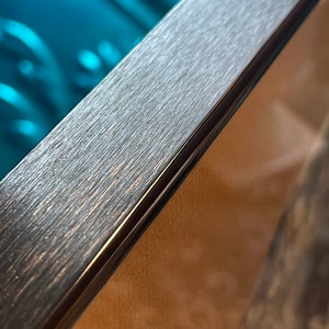 May include: Close-up of a dark gray picture frame with a brushed metal finish. The frame has a thin black border.