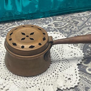 May include: A small, round copper pot with a matching lid featuring a series of diamond-shaped cutouts. The pot has a single, long, slightly curved handle made of dark wood, which includes a small loop of twine attached to the end for hanging.