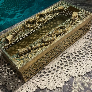 May include: Gold metal tissue box cover with ornate design and tassel accents. The cover has a clear plastic window to view the tissues.