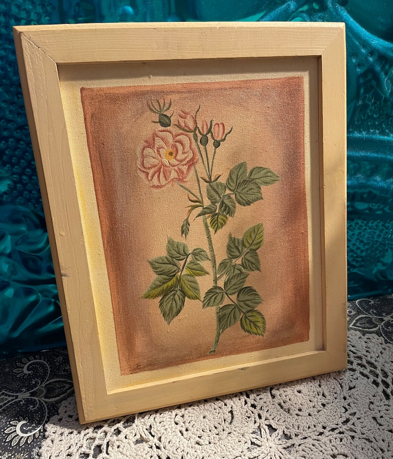 Puede incluir: Una pintura enmarcada de una sola rosa rosa con hojas verdes y capullos sobre un fondo beige. El marco es de madera beige claro.