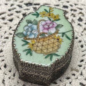 May include: A small, green porcelain trinket box with a silver metal rim. The lid features a hand-painted design of a basket of flowers with yellow, pink, and blue flowers.