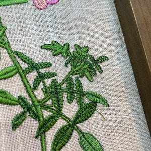 May include: Embroidered floral design on a beige linen fabric with a wooden frame. The design features a pink flower with green leaves and stems.