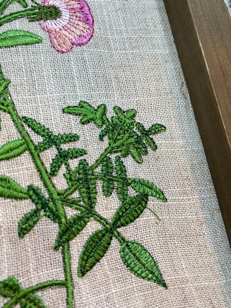 May include: Embroidered floral design on a beige linen fabric with a wooden frame. The design features a pink flower with green leaves and stems.