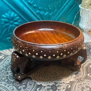 May include: A wooden bowl with intricate inlaid designs sits on a stand with four elephant legs. The bowl is dark brown with a light brown interior and features a decorative border of white and gold accents.