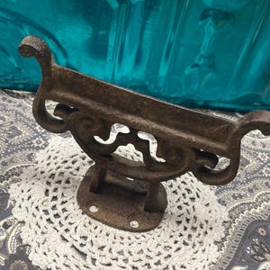 May include: A rustic, brown cast iron wall-mounted shelf with ornate scrollwork and a curved top. The shelf is mounted on a small, rectangular base with two screw holes. It is sitting on a white crocheted doily.