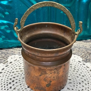 May include: A small copper bucket with a brass handle. The bucket has a hammered finish and is slightly tarnished. The handle is decorated with a geometric pattern.