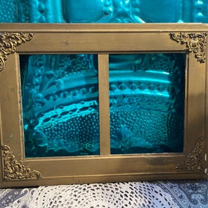 K&ouml;nnte beinhalten: Ein goldfarbener Bilderrahmen mit verzierten Details und zwei Glasscheiben. Der Rahmen ist aus Holz und hat eine patinierte Oberfl&auml;che.