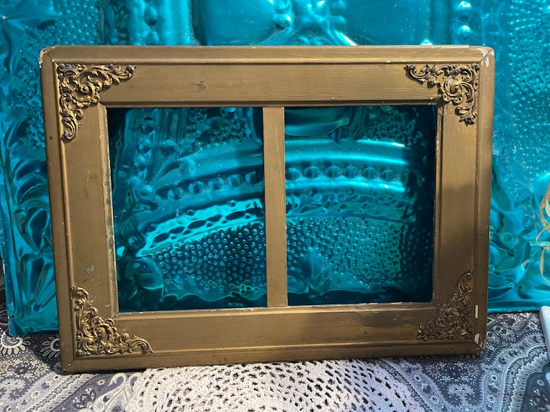 K&ouml;nnte beinhalten: Ein goldfarbener Bilderrahmen mit verzierten Details und zwei Glasscheiben. Der Rahmen ist aus Holz und hat eine patinierte Oberfl&auml;che.