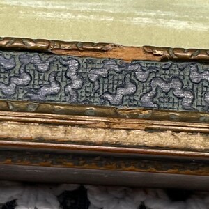 May include: Close-up of a wooden picture frame with a gold-colored finish. The frame has a decorative border with a repeating pattern of small, dark green shapes. The frame is slightly damaged, with a small chip in the wood.