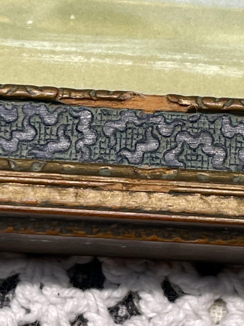 May include: Close-up of a wooden picture frame with a gold-colored finish. The frame has a decorative border with a repeating pattern of small, dark green shapes. The frame is slightly damaged, with a small chip in the wood.