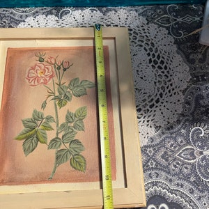 Puede incluir: Una pintura enmarcada de una sola rosa rosa con hojas y tallos verdes sobre un fondo rosa claro. El marco es de madera clara.