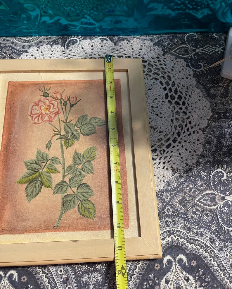 Puede incluir: Una pintura enmarcada de una sola rosa rosa con hojas y tallos verdes sobre un fondo rosa claro. El marco es de madera clara.