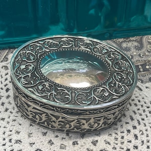 May include: An oval, silver-toned trinket box with ornate floral and scrollwork detailing. The lid features a clear, oval window, revealing the interior. The box sits on a white, crocheted surface, with a teal background.