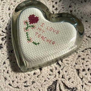 May include: A clear glass heart-shaped paperweight. Inside, a cross-stitched design features a red heart and flowers with the words "I LOVE MY TEACHER" in red thread. The paperweight is on a white crocheted surface.