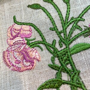May include: Embroidered floral design on a beige linen fabric. The design features a pink flower with green stems and leaves.