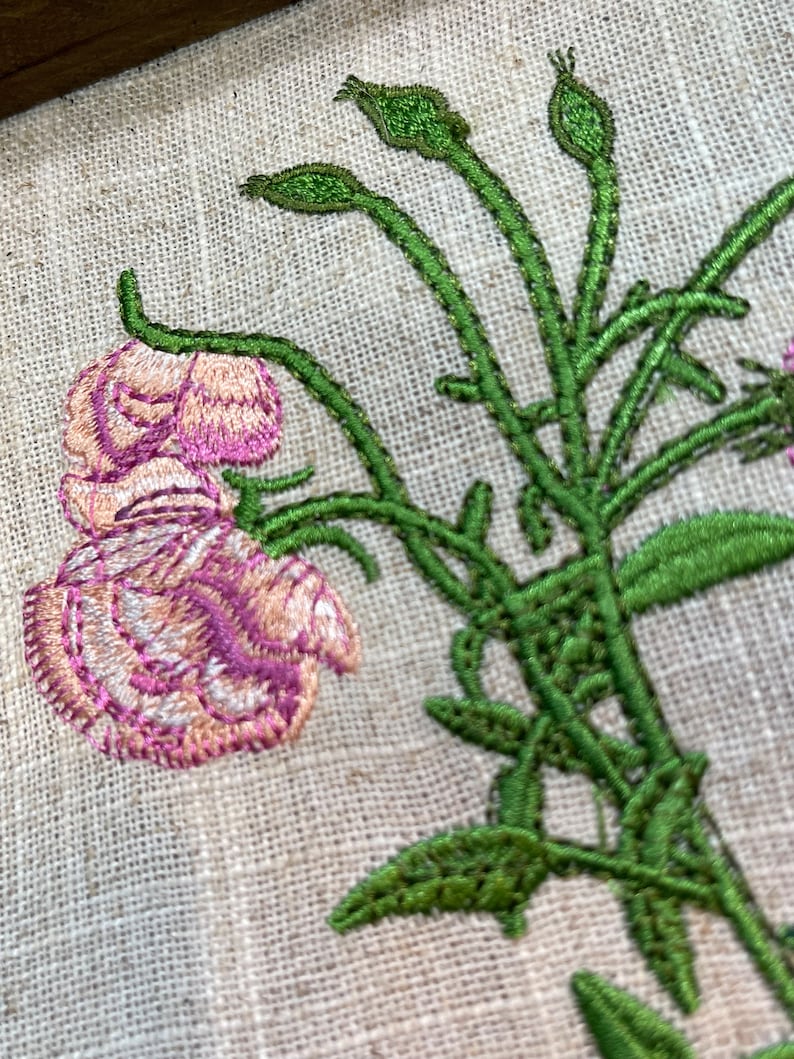 May include: Embroidered floral design on a beige linen fabric. The design features a pink flower with green stems and leaves.