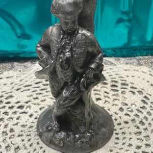 May include: A silver-colored figurine of a man in period clothing, holding a hat. The figure is mounted on a round base. The figurine appears to be made of metal and has intricate details in the clothing and facial features.