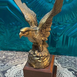 May include: A bronze-colored eagle sculpture with outstretched wings, perched atop a rocky base and a dark wooden pedestal. The eagle's feathers are detailed, and the sculpture has a metallic sheen. The background is a teal-colored surface.