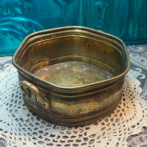 May include: A vintage brass planter with a hexagonal shape and a hammered finish. The planter has a single handle on one side and is slightly tarnished.