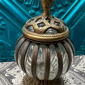 May include: An antique glass jar with a decorative brass lid and base. The jar has a clear, ribbed glass body with brass accents. The lid features a geometric design and a small finial. The jar sits on a white crocheted doily.