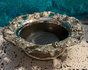 May include: Silver wine bottle holder with a grapevine design. The holder has a round base and a raised rim.