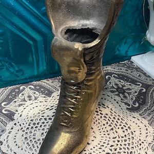 May include: A gold-colored metal boot-shaped vase with a lace-up design. The vase is sitting on a white crocheted doily.