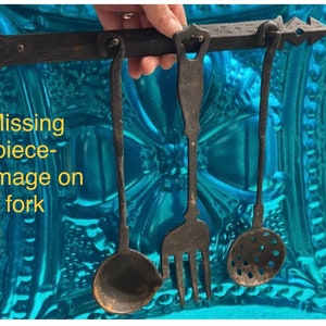 May include: A set of three antique kitchen utensils hanging on a wooden rack. The utensils include a ladle, a fork, and a slotted spoon. The fork has a missing piece, showing signs of damage.