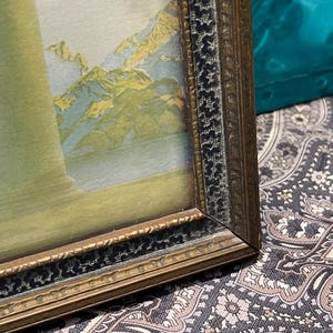 May include: A framed picture with a landscape scene of mountains and water. The frame is gold with a black inner border.