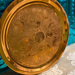 May include: A gold-colored metal tray with an ornate, embossed design. The tray has a raised rim and is slightly worn, indicating age. The text "Aged!" is visible in the top left corner of the image.