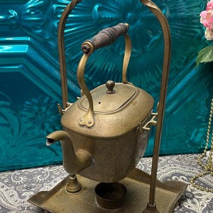 May include: Antique brass kettle with a hammered finish, featuring a wooden handle and a spout. The kettle sits on a matching tray with a stand. The design includes a decorative arched handle. A vintage tea kettle.