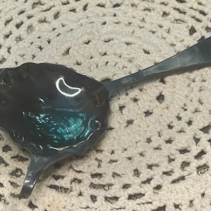 May include: A silver tea strainer with a decorative, scalloped edge and a long handle. The strainer is sitting on a white crocheted doily.