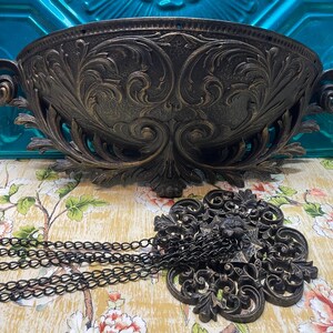 May include: Ornate, dark bronze-colored wall planter with intricate floral and scrollwork details. Below, a matching decorative medallion with a chain. The planter has two handles and a wide, curved bowl shape. The items are on a floral patterned surface.