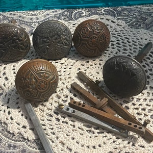May include: A collection of antique bronze door knobs with intricate embossed designs. The knobs vary in size and pattern, showcasing floral and geometric motifs. Some knobs are detached, with their spindles and hardware visible.