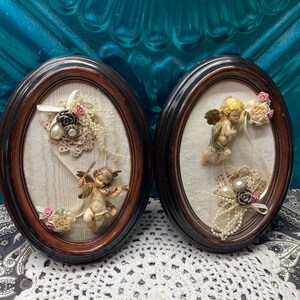 May include: Two oval wall frames with dark wood borders containing cream fabric backings. Each frame features a three-dimensional cherub figurine, delicate lace accents, faux pearl strands, and small fabric flowers arranged in a decorative display.