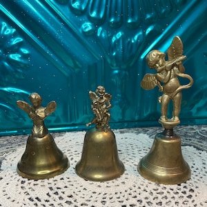 May include: Three antique brass bells with angel figurines. The bells vary in size and design, with one angel playing a flute. The bells are displayed on a white lace doily against a teal background.