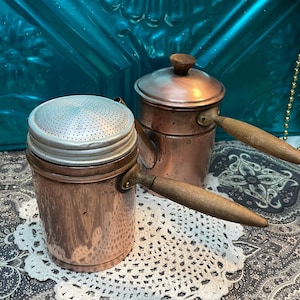 May include: Two antique copper coffee pots with wooden handles. One pot has a perforated lid. The pots are cylindrical with a shiny, aged finish. The handles are attached with brass fittings. The background is a teal-colored surface.
