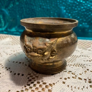 May include: A small, round, brass incense burner with a decorative design of a mythical creature on the side. The burner has a slightly flared rim and a rounded base.