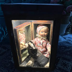 May include: A vintage doll with a pink and blue dress is displayed in a wooden box with a glass front. The doll is wearing a white bonnet and has a black shoe.