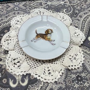 May include: White ceramic ashtray with a gold rim and a running dog illustration. The ashtray is placed on a white, crocheted doily. The background features a patterned fabric with shades of gray and white.