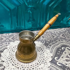 May include: A brass Turkish coffee pot with a wooden handle. The pot has a hammered finish and a small spout. The handle is brown with black stripes.