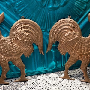 May include: Two copper rooster wall decor pieces. The roosters are facing each other with their wings spread out. They have a detailed design with a textured surface.
