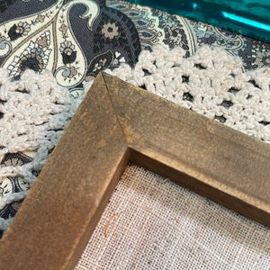 May include: Close-up of the corner of a brown wooden frame surrounding a piece of beige linen fabric. The frame is made of wood with a dark brown stain.
