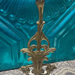May include: Ornate brass candlestick holder with a detailed floral design. The candlestick has a square base and a tall, slender stem with decorative flourishes. The top cup is designed to hold a single candle. The candlestick is approximately 8 inches tall.