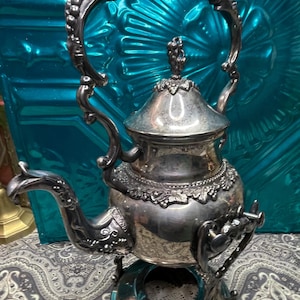 May include: Ornate silver tea kettle with a decorative handle and lid finial. The kettle sits on a matching stand with intricate detailing. The design features floral and scrollwork patterns, suggesting a vintage or antique style. The background is a teal-colored surface.