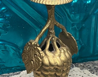 May include: A gold-toned brass candlestick shaped like a bunch of grapes. The candlestick has a round base with intricate leaf details and a cup-shaped top for holding a candle.