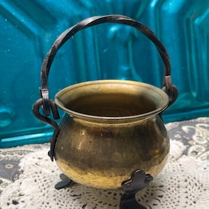May include: A small brass cauldron with a rounded body, a wide opening, and a dark metal twisted handle. The vessel is supported by three dark metal feet attached to the base, featuring a polished brass finish with visible signs of age.