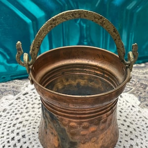 May include: A small copper bucket with a hammered finish and a brass handle. The bucket has a worn, aged look.