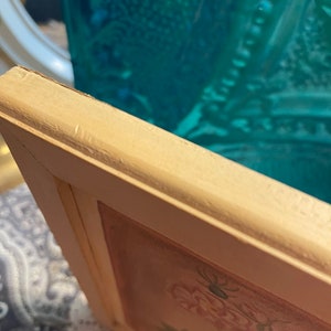 Puede incluir: Primer plano de un marco de imagen de madera blanca con un dise&ntilde;o floral en el interior. El marco tiene un aspecto desgastado y est&aacute; ligeramente astillado en el borde.