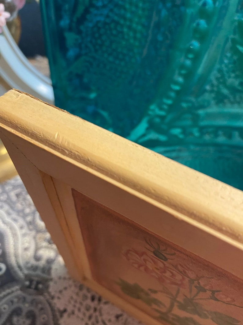 Puede incluir: Primer plano de un marco de imagen de madera blanca con un dise&ntilde;o floral en el interior. El marco tiene un aspecto desgastado y est&aacute; ligeramente astillado en el borde.